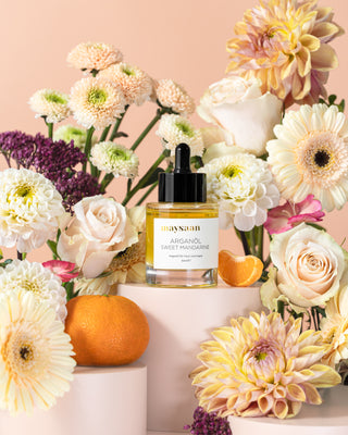 maysaan Arganöl Sweet Mandarine – Flasche zwischen hellen Blüten und Mandarinen auf pastellfarbenem Hintergrund. Pflegeöl aus Arganöl für Haut und Haare.