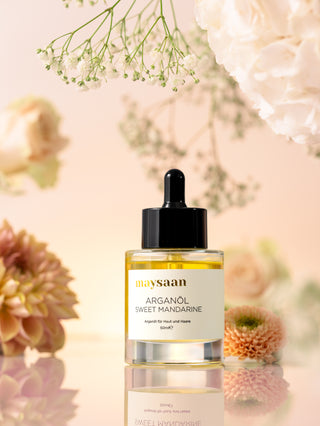 maysaan Arganöl Sweet Mandarine – Glasflasche umgeben von zarten Blumen, natürliche Produktinszenierung. Pflegeöl aus Arganöl für Haut und Haare.
